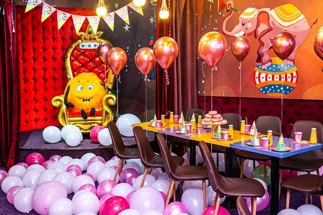 Sala Circus decorată pentru o sărbătoare: un tron auriu cu un personaj galben, masa aranjată, o ghirlandă și baloane roz și albe.