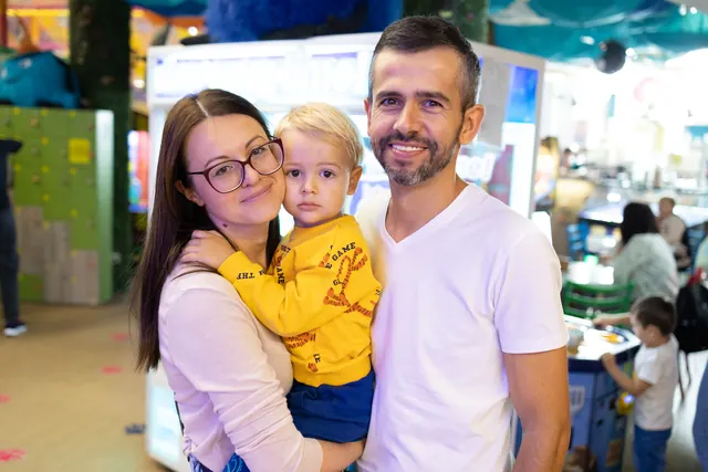 O femeie și un bărbat care ține un copil în brațe zâmbesc în fața aparatelor de joc.