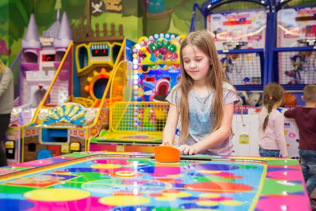 Un copil cu un mâner portocaliu se joacă la o masă de air hockey multicoloră.