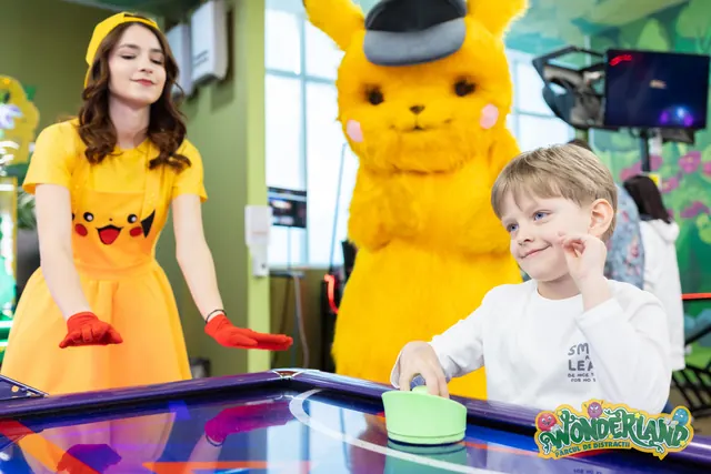 Un copil zâmbește la masa de air hockey, lângă o femeie într-o rochie galbenă și un personaj costumat galben.